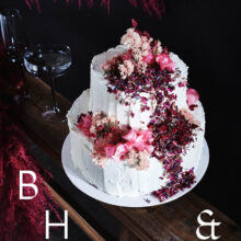 Food photography of white and pink wedding cake topped with pink raspberries and flowers in front of black background