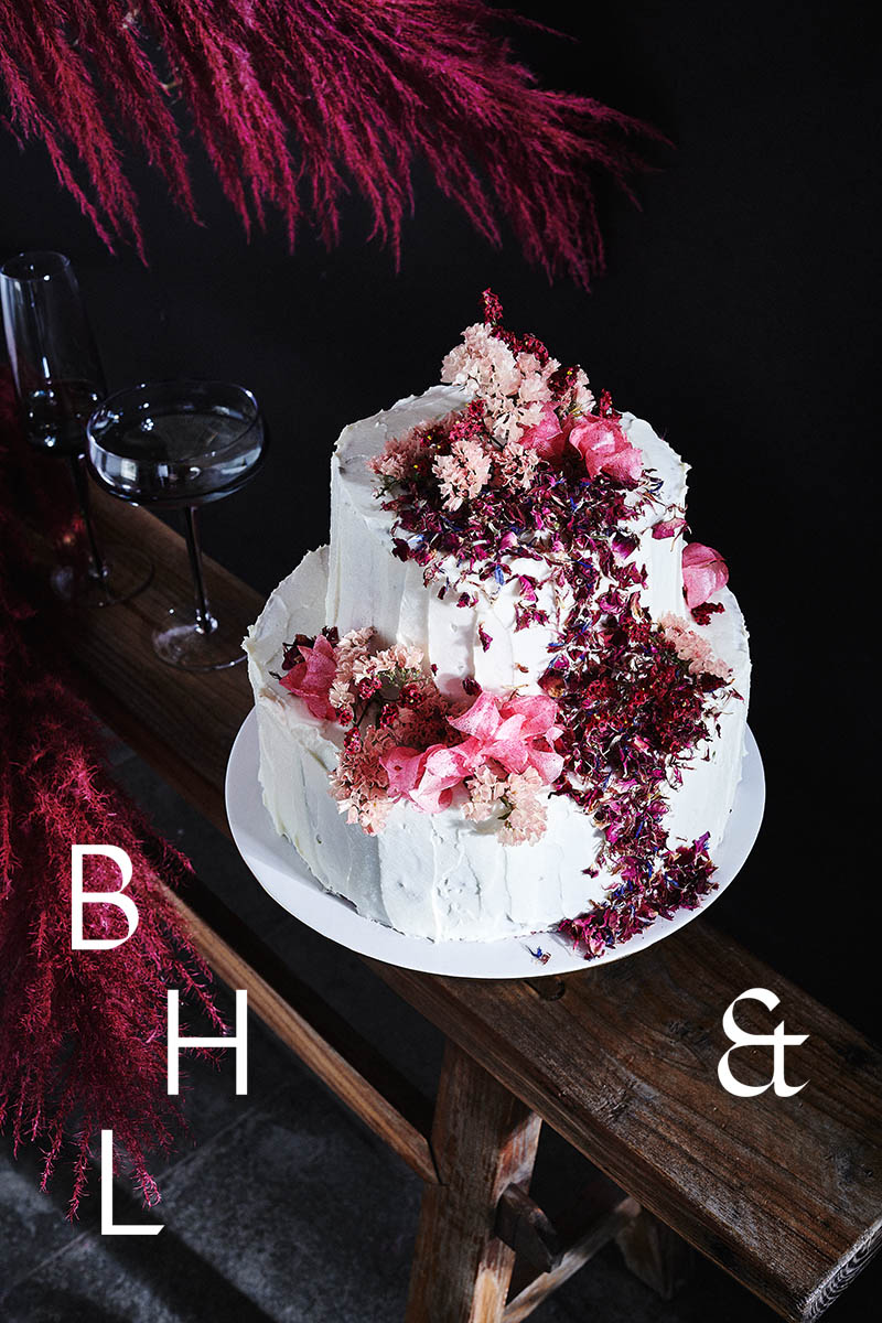 Food photography of white and pink wedding cake topped with pink raspberries and flowers in front of black background