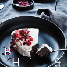 Food photography of white and pink cake with chocolate and dried berries in a black table setting with dark linen and black tableware decorated with dry bamboo in scandinavian industrial loft