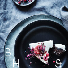 Food photography of white and pink cake with chocolate and dried berries in a black table setting with dark linen and black tableware decorated with dry bamboo in scandinavian industrial loft