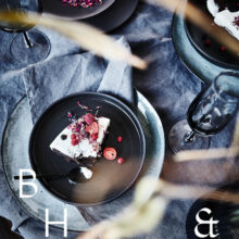 Food photography of white and pink cake with chocolate and dried berries in a black table setting with dark linen and black tableware decorated with dry bamboo in scandinavian industrial loft